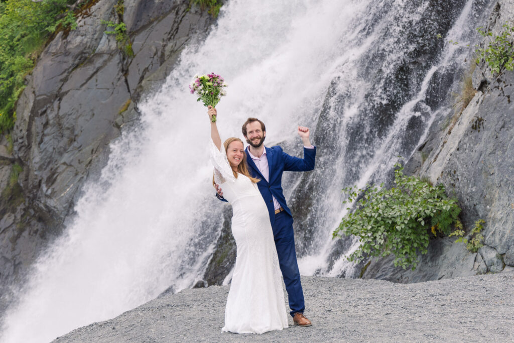 Bride and groom celebrate in front of waterfall during elopement in Seward, Alaska