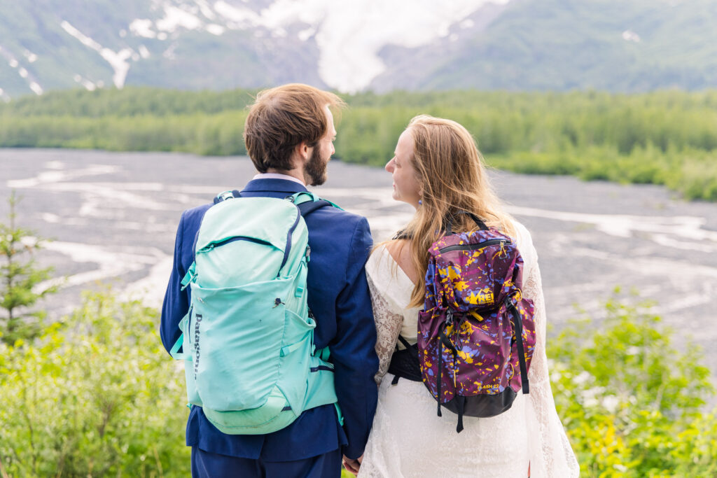 Wedding couple smiles in front of Exit Glacier with adventure hiking backpacks during Alaska elopement