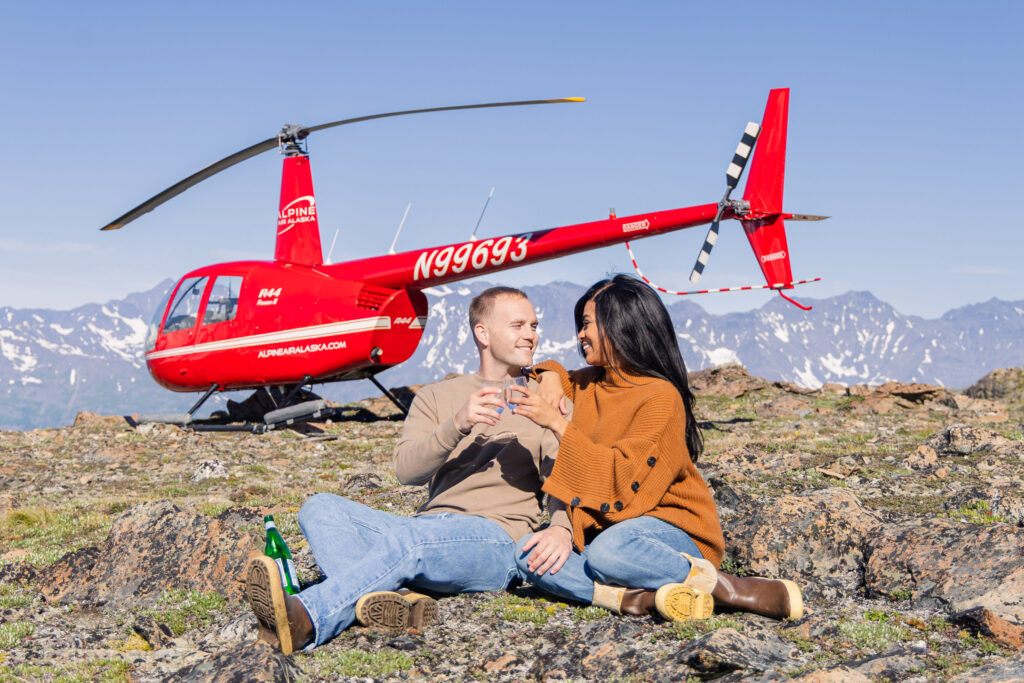 Recently engaged couple drinks champagne in front of a helicopter while overlooking a glacier in Alaska. Their proposal was captured by Aronoff Photography minutes before this picture.