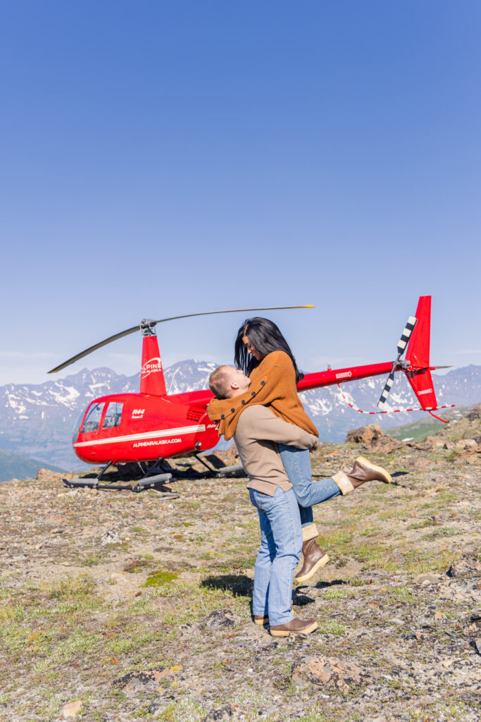 Man lifts fiancé in romantic pose after getting engaged in front of helicopter and glacier in Alaska.