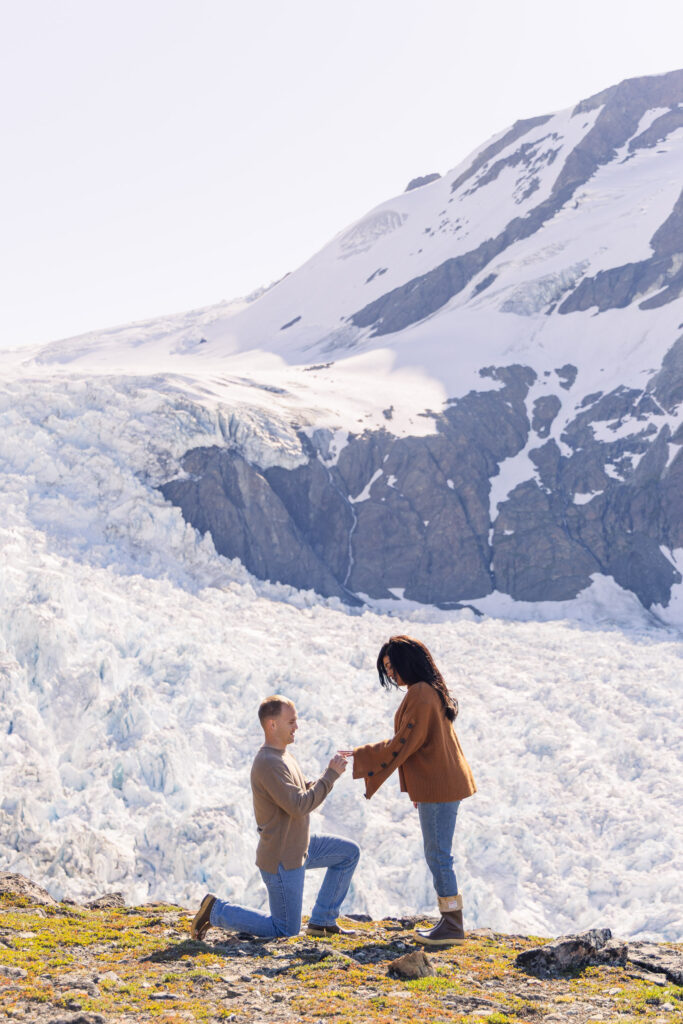 Man kneels down and proposes to girlfriend in front of glacier in Alaska.