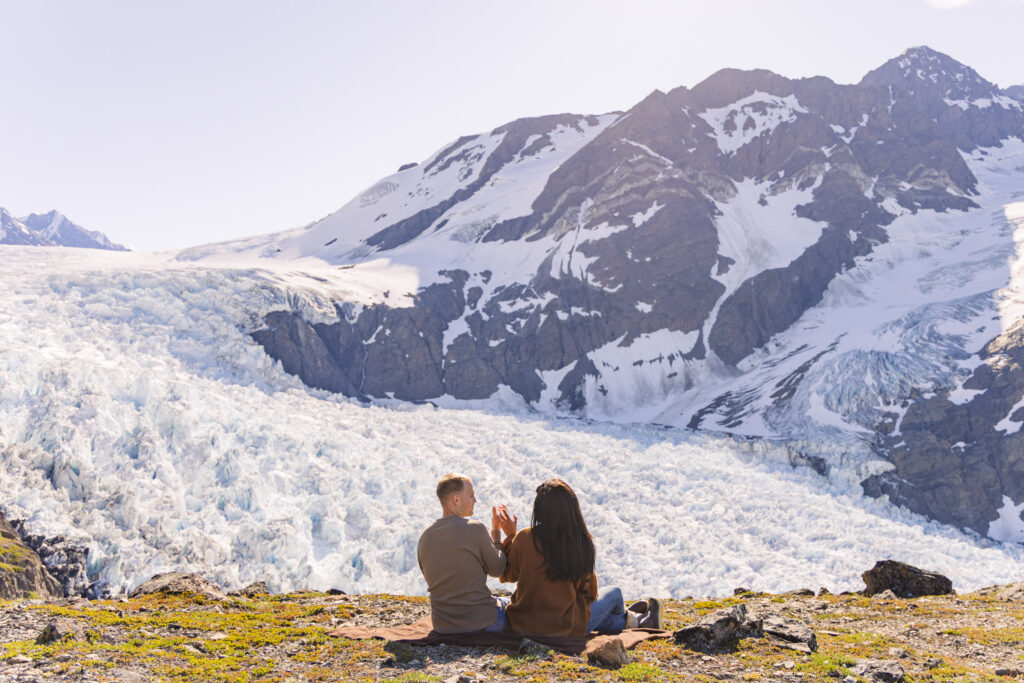 Recently engaged couple has picnic overlooking Alaskan glacier on cliff after helicopter landing.