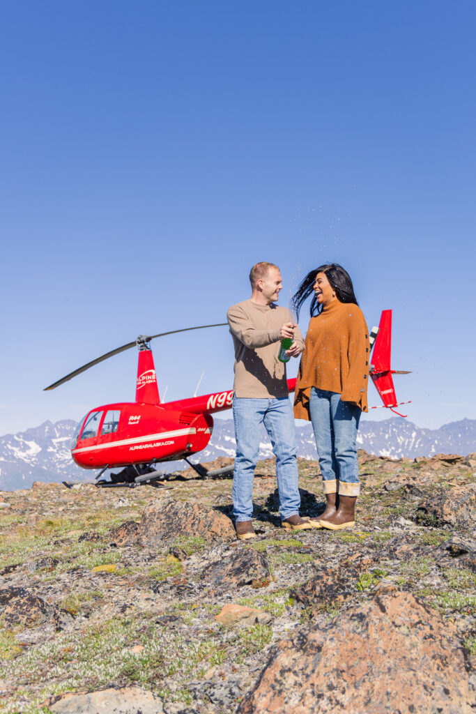 Couple pops champagne in front of a helicopter in Alaska.