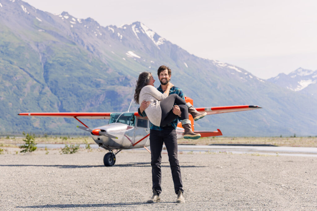 Couple holds each other in front of their Cessna 172 airplane while taking couple's portraits on a landing strip in the Alaskan backcountry