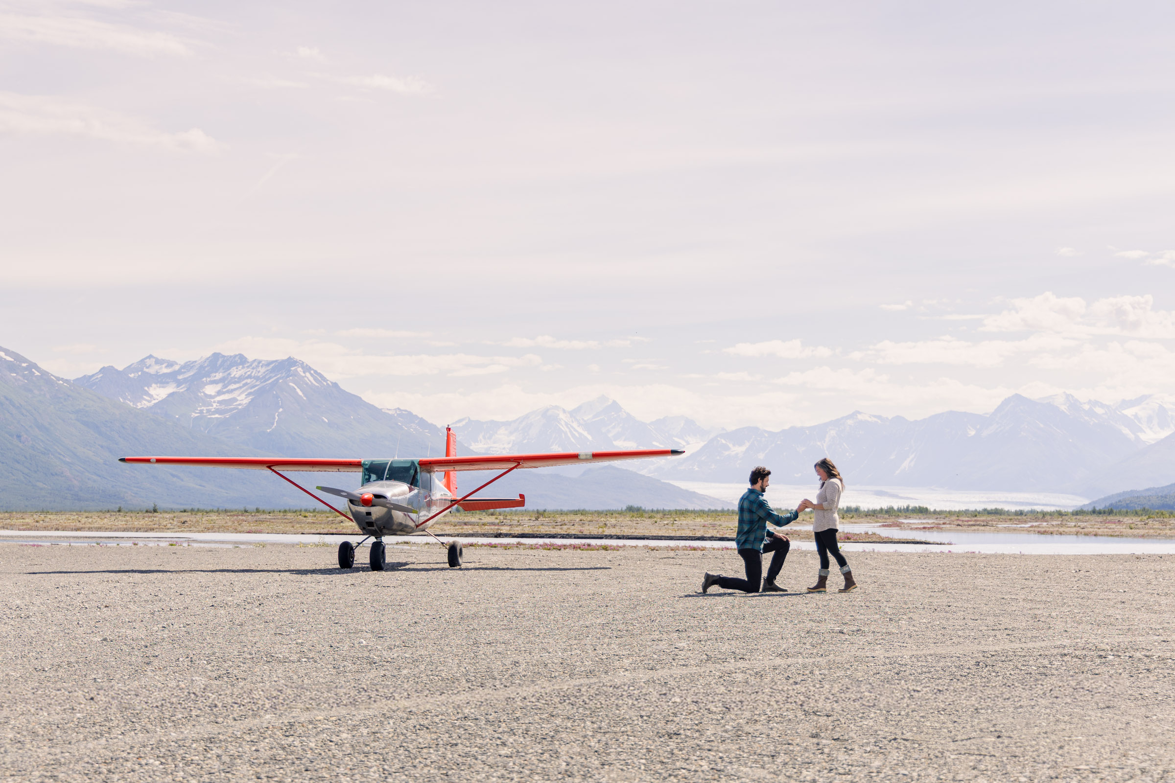 Man proposes to girlfriend in Alaska backcountry, in front of a Cessna airplane, mountains, and a glacier.