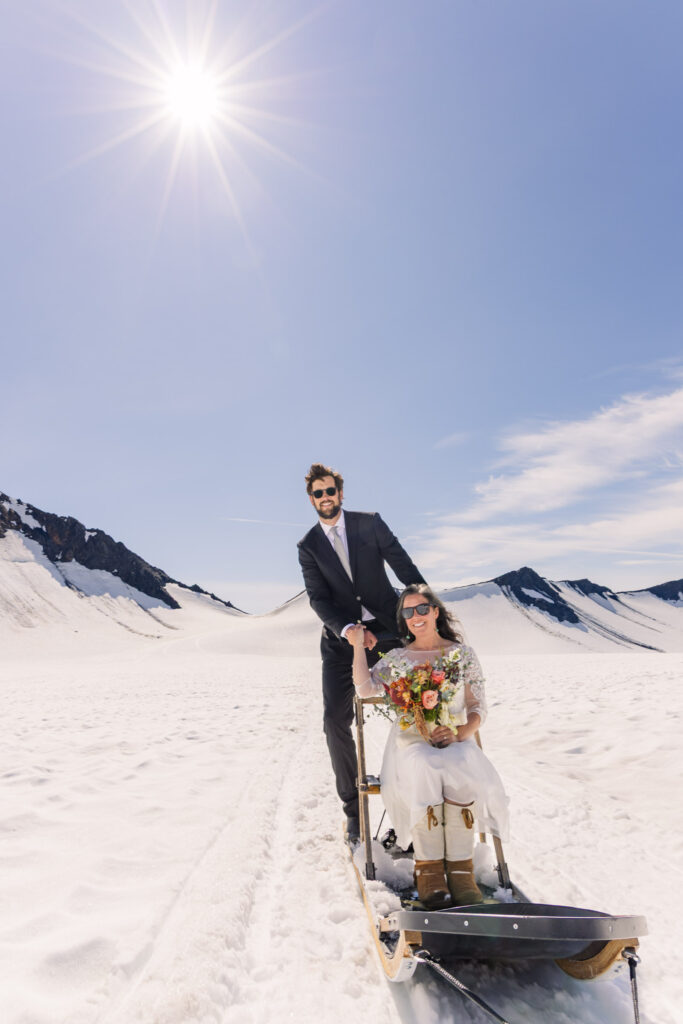 Groom mushing dogsled team while bride rides in dogsled during Alaska dogsledding glacier elopement