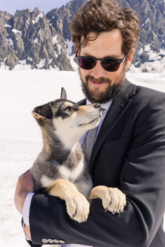 Groom playing with husky puppy at glacier dogsled camp during Alaska elopement