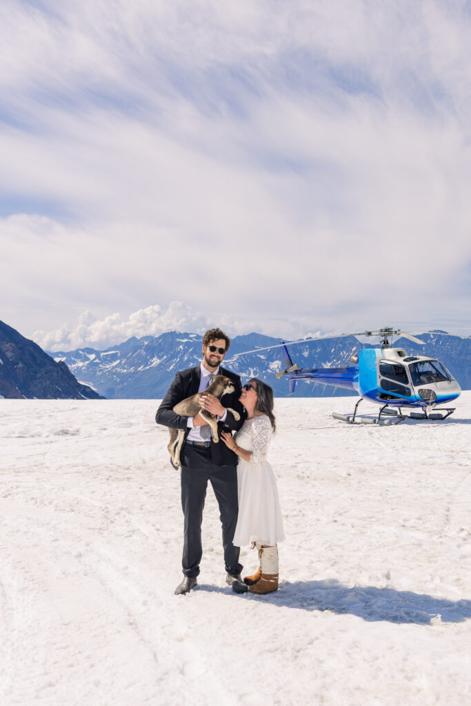 Couple playing with husky puppy in front of helicopter at glacier dogsled camp during Alaska elopement