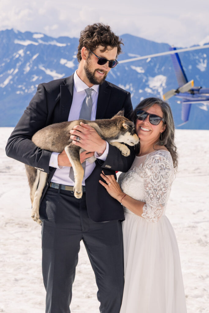 Couple playing with husky puppy in front of helicopter at glacier dogsled camp during Alaska elopement