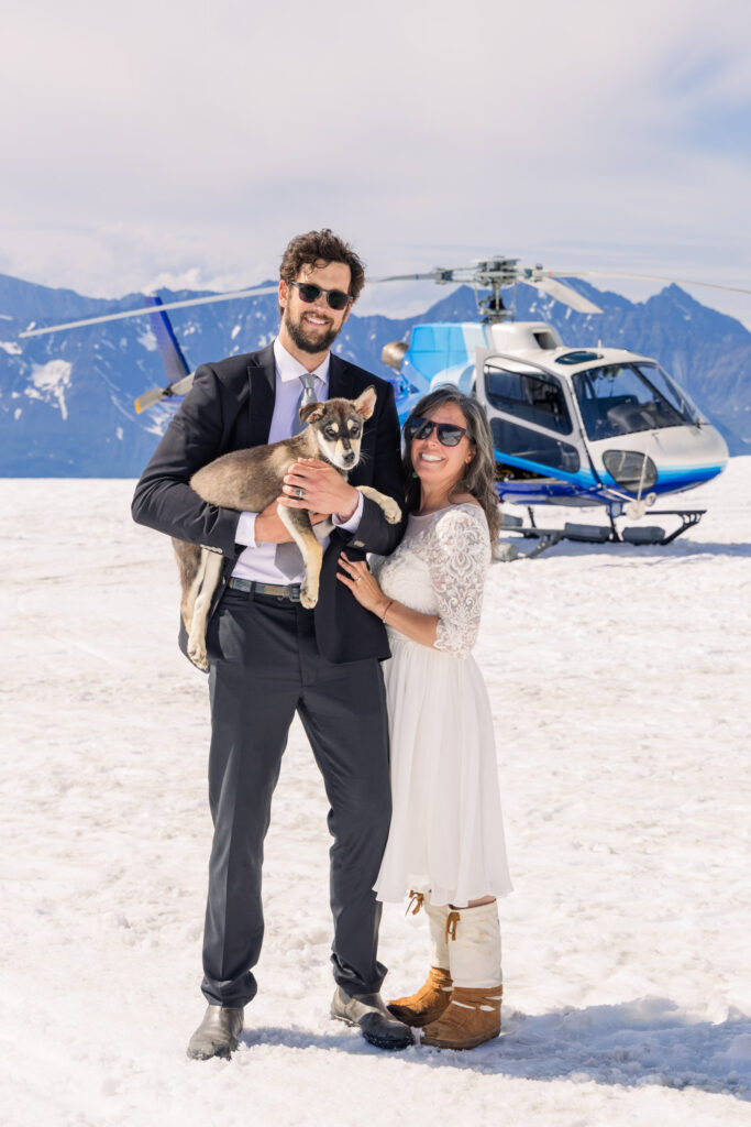 Couple playing with husky puppy in front of helicopter at glacier dogsled camp during Alaska elopement