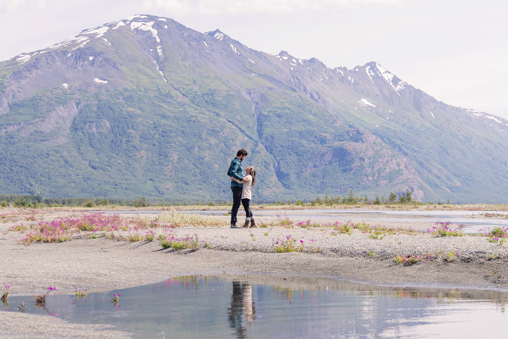 Couple gazes lovingly at each other during engagement/proposal portraits in the Alaskan backcountry, with a small lake in the foreground and large mountain in the background