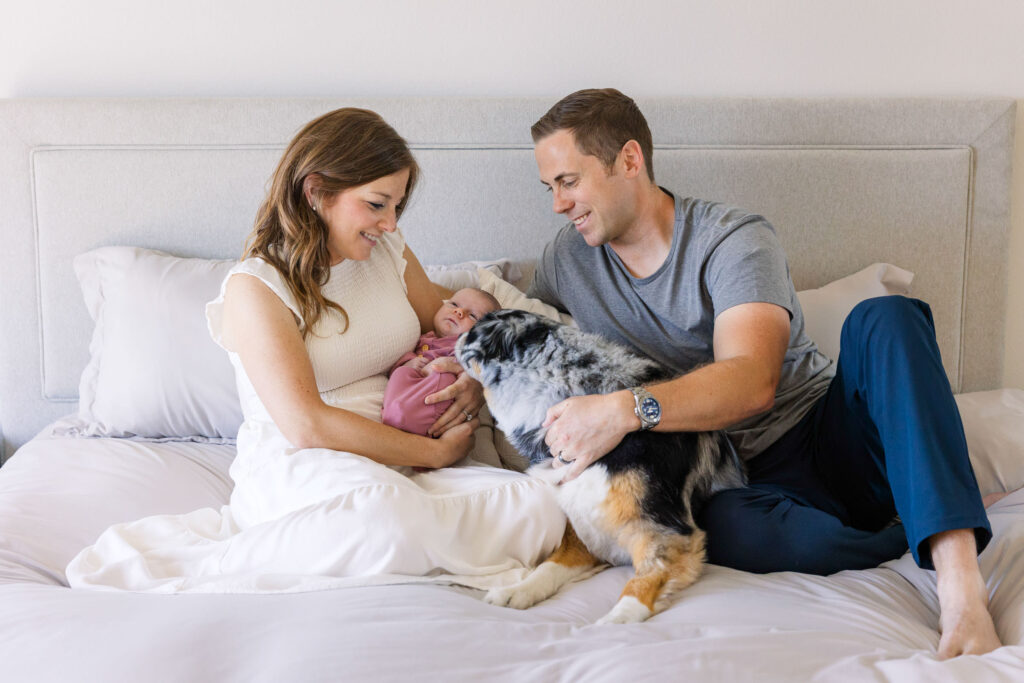 parents sitting on bed with newborn and dog interacting with the baby