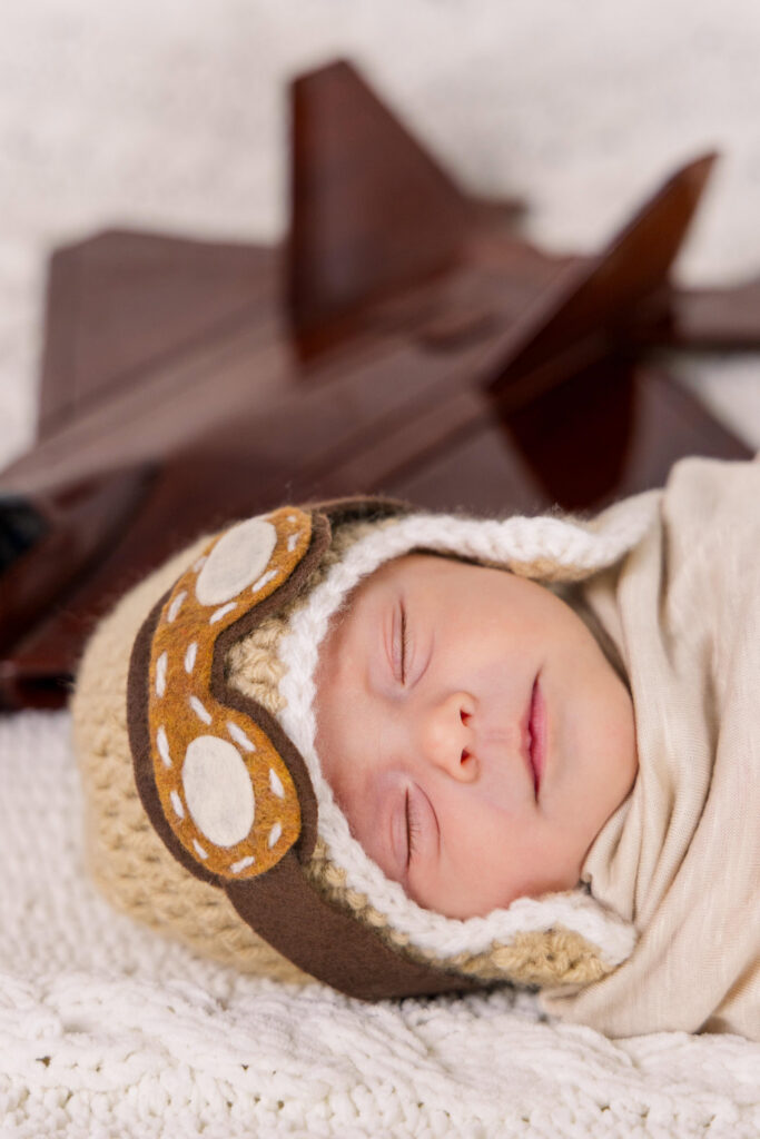 newborn wearing knit pilot cap and goggles in front of model f-22 aircraft