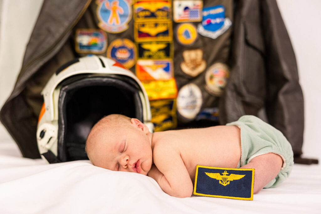 newborn in front of a navy pilot helmet and flight jacket with patches