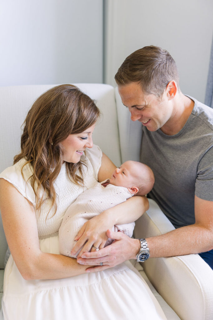 mother holding newborn in nursing chair while father kneels next to them and smiles at baby