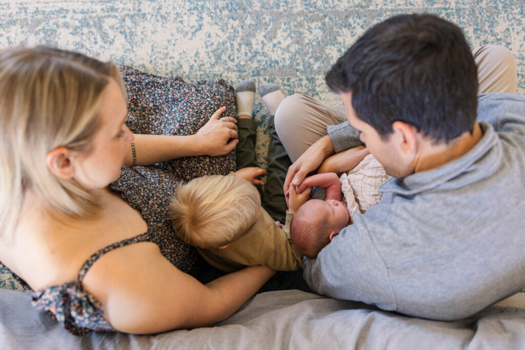 parents looking down at newborn baby they are holding with toddler holding baby's hand during lifestyle newborn session