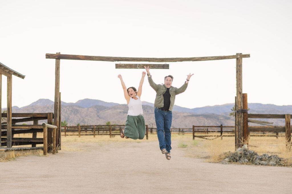 David and Lisa jump for joy in front of corral and mountains after getting engaged in historic Pioneertown.
