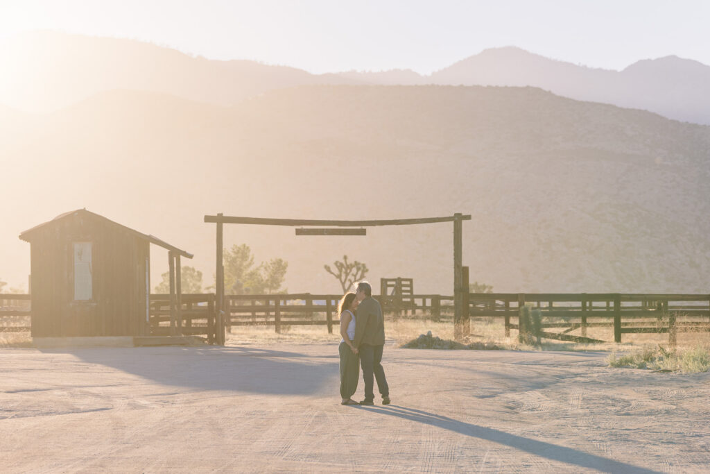 David and Lisa embracing in the golden evening light near Pioneertown, with an old corral, Joshua trees, and desert mountains in the background.